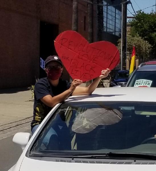 Jack in a car caravan in 2020, wearing a mask and holding a sign that says "Revolutionary Love for Black Lives"